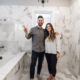 A bathroom with a newly renovated shower stall featuring large tiles, a rainfall showerhead, and a glass door. The vanity has a modern design with a white countertop and sleek metal hardware. The bathroom also features a new toilet and fresh coat of paint on the walls.